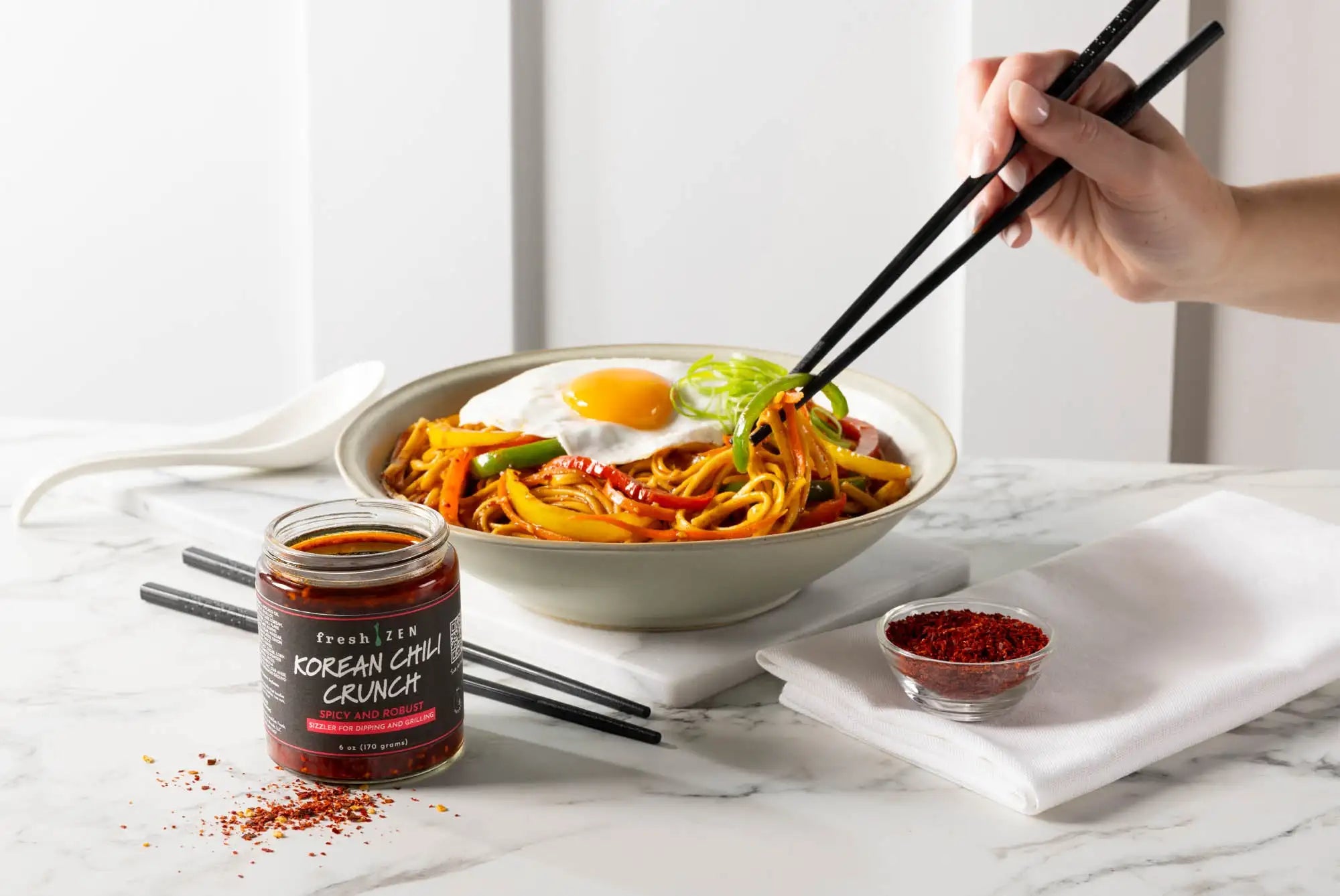 Bowl of noodles with a fried egg, garnished with green onions, on a marble surface with a jar of Korean chili crunch seasoning.