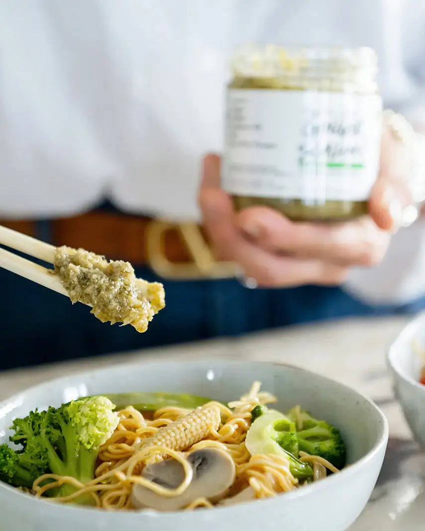 A jar of green pesto with a white label, held in hands above a bowl of noodles and vegetables.