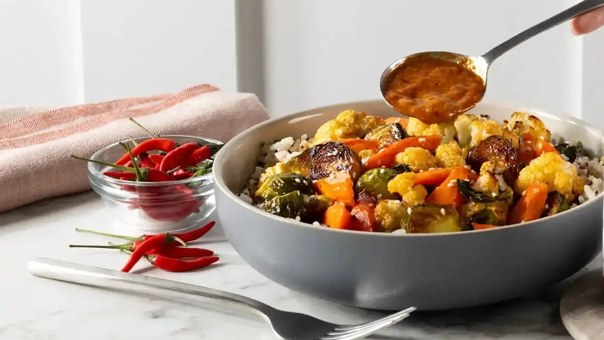 A gray ceramic bowl filled with a colorful mix of roasted vegetables and rice, topped with a spoonful of orange sauce being poured over it.