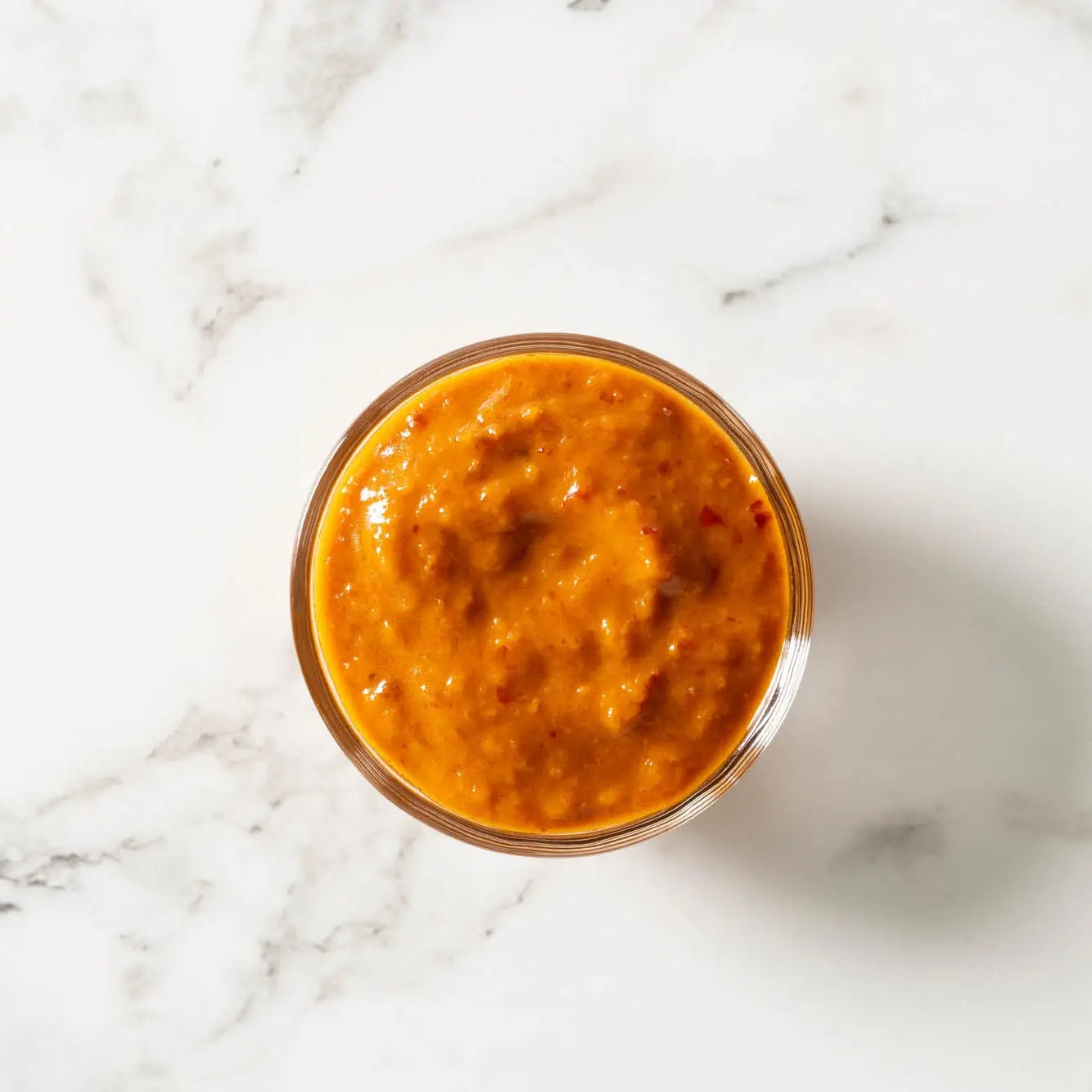 A clear glass jar of thai chili garlic sauce with vibrant orange-red color and visible red pepper flecks