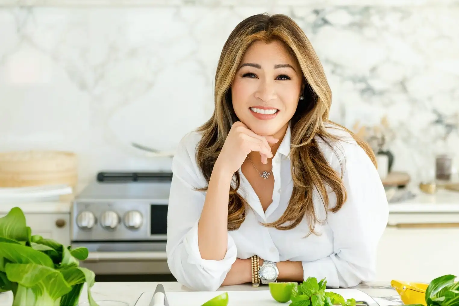 A woman with long, wavy brown hair wearing a white button-up shirt and a silver necklace.